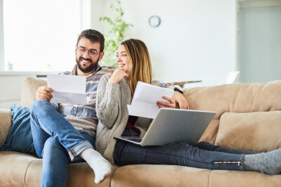 Couple at home on the couch looks at paperwork and laptop to consider the pros and cons of refinancing their home.